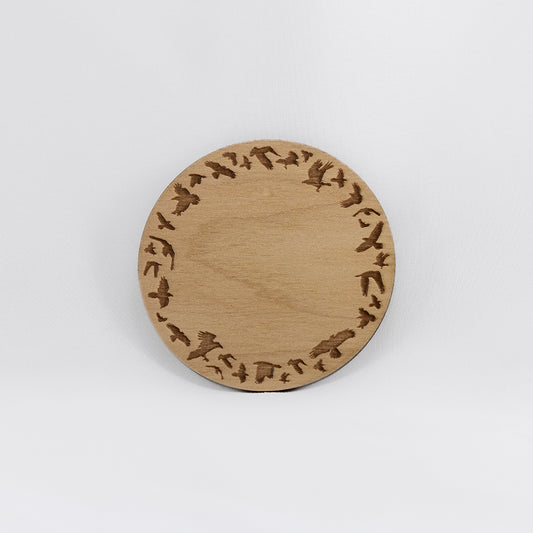 A circular wooden laser-cut crystal grid engraved with a border made up of ravens in flight with a plain white background. 