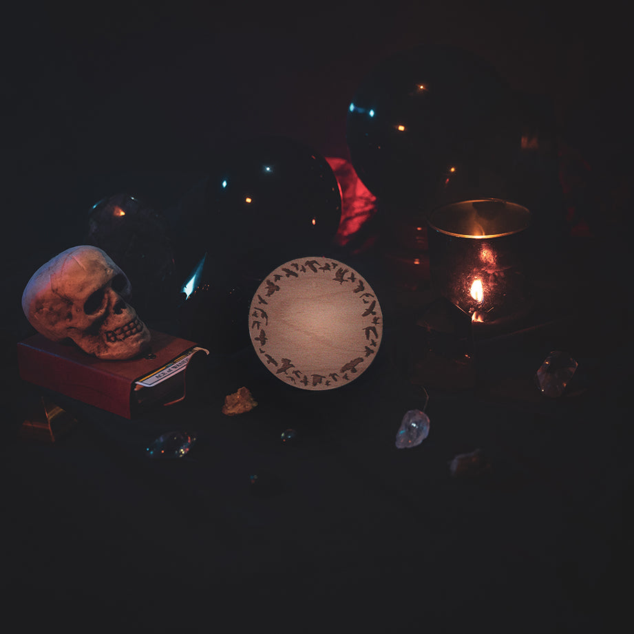 A circular wooden laser-cut crystal grid engraved with a border made up of ravens in flight. The grid sits amongst candles, crystal balls, a skull, tarot cards and assorted crystals.