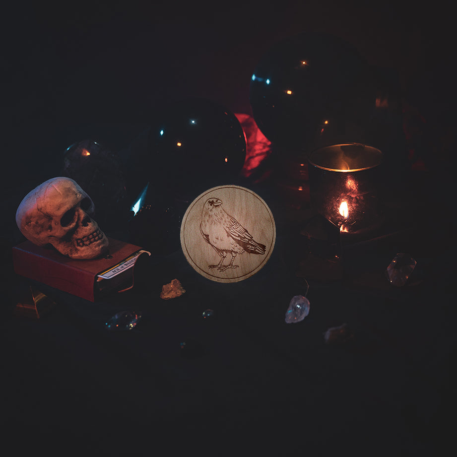 A circular wooden laser-cut crystal grid engraved with a raven. The grid sits amongst candles, crystal balls, a skull, tarot cards and assorted crystals.