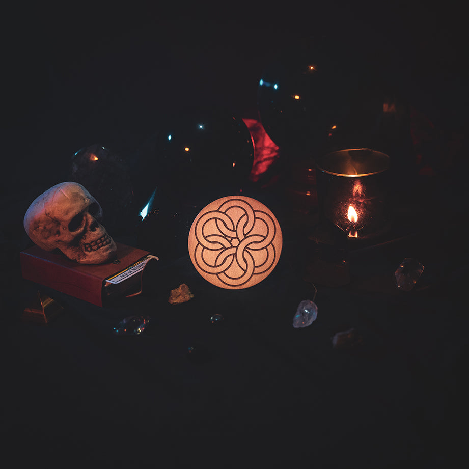 A circular wooden laser-cut crystal grid engraved with a Celtic ring knot. The grid sits amongst candles, crystal balls, a skull, tarot cards and assorted crystals.