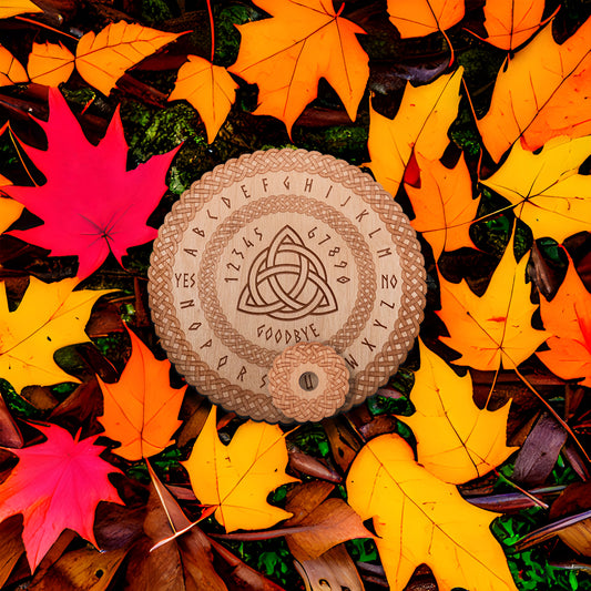 Circular Baltic birch laser-cut spirit  board engraved with the words "yes", "no", "goodbye", as well as the entire alphabet lays on the leaf covered ground. The board has the Celtic Triquetra symbol in the centre and a decorative die-cut braid border. A small circular planchette with a die-cut edge sits on top of the board.