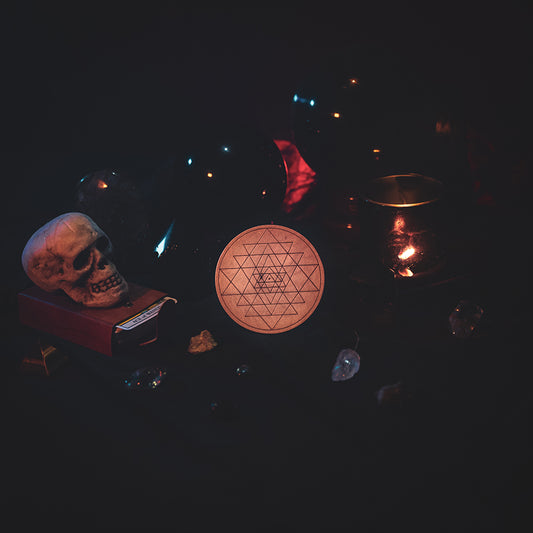 A circular wooden laser-cut crystal grid engraved with Sri Yantra. The grid sits amongst candles, crystal balls, a skull, tarot cards and assorted crystals.