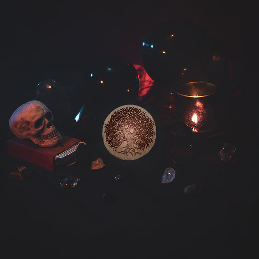 A circular wooden laser-cut crystal grid engraved with a gnarly old tree. The grid sits amongst candles, crystal balls, a skull, tarot cards and assorted crystals.