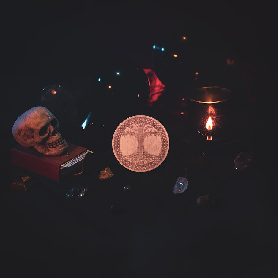 A circular wooden laser-cut crystal grid engraved with the Celtic Tree of Life. The grid sits amongst candles, crystal balls, a skull, tarot cards and assorted crystals.