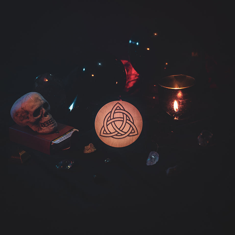 A circular wooden laser-cut crystal grid engraved with the Celtic Triquetra symbol. The grid sits amongst candles, crystal balls, a skull, tarot cards and assorted crystals.