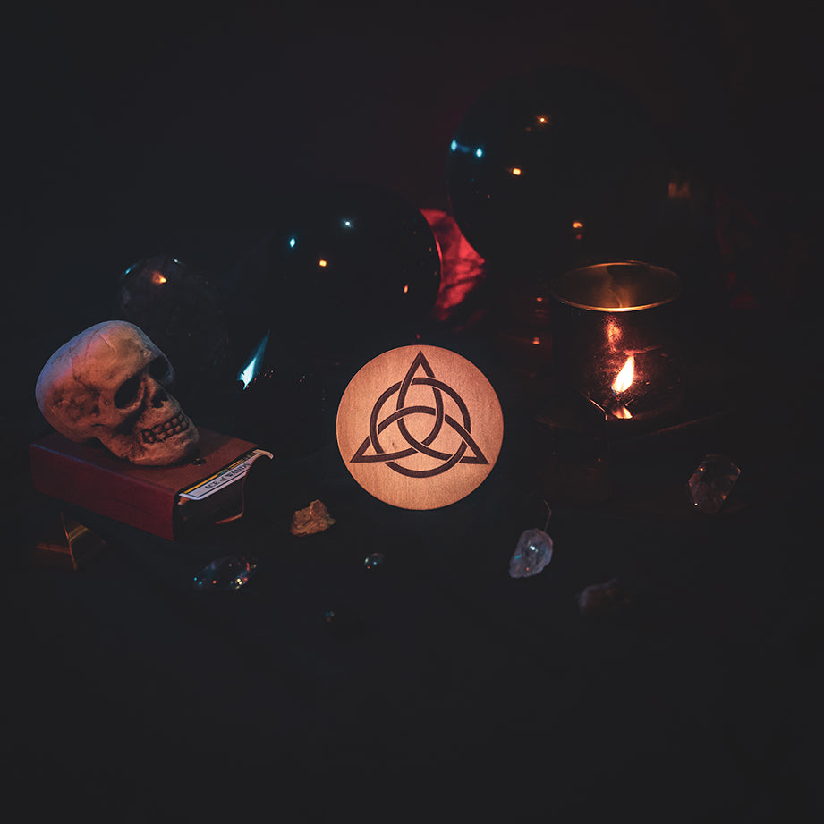 A circular wooden laser-cut crystal grid engraved with the Celtic Triquetra symbol. The grid sits amongst candles, crystal balls, a skull, tarot cards and assorted crystals.