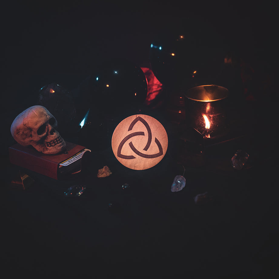 A circular wooden laser-cut crystal grid engraved with the Celtic Triquetra symbol. The grid sits amongst candles, crystal balls, a skull, tarot cards and assorted crystals.