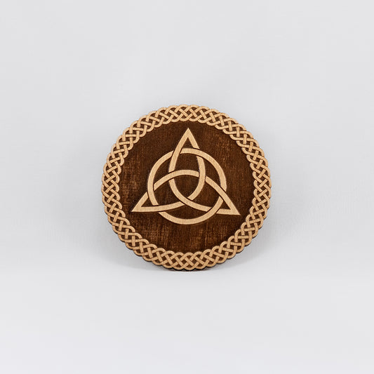 A circular wooden laser-cut crystal grid engraved with the Celtic Triquetra symbol and a scalloped braid border with a plain white background. 