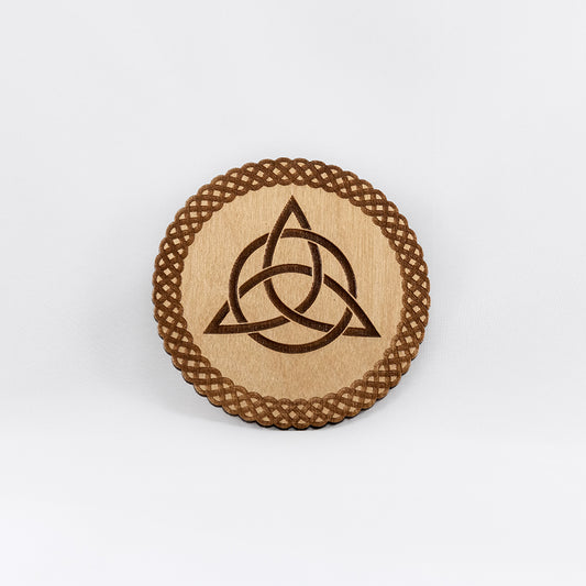 A circular wooden laser-cut crystal grid engraved with the Celtic Triquetra symbol and a scalloped braid border with a plain white background. 