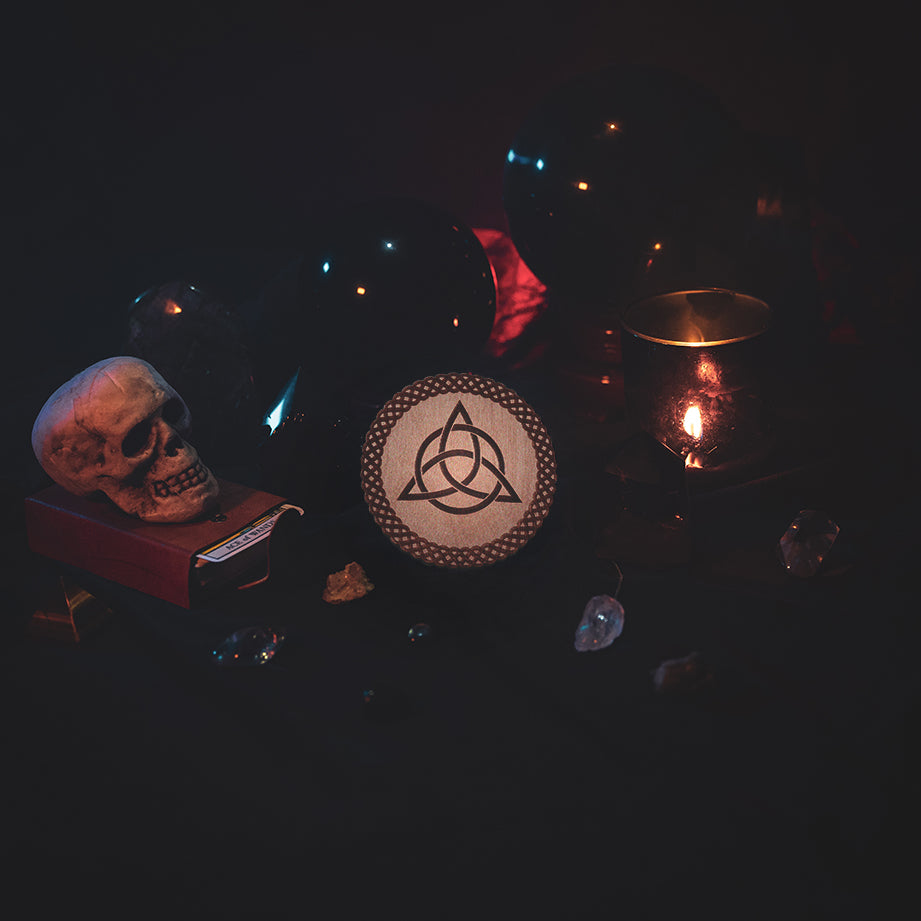 A circular wooden laser-cut crystal grid engraved with the Celtic Triquetra symbol and a scalloped braid border. The grid sits amongst candles, crystal balls, a skull, tarot cards and assorted crystals.