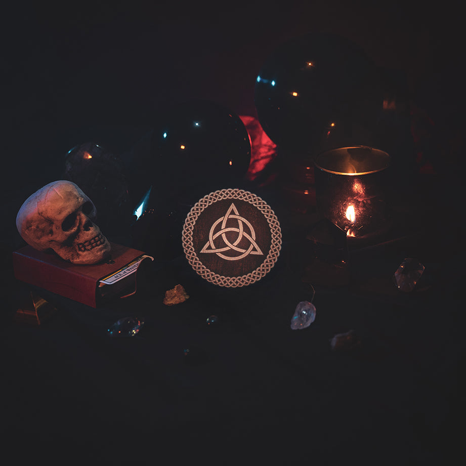 A circular wooden laser-cut crystal grid engraved with the Celtic Triquetra symbol and a scalloped braid border. The grid sits amongst candles, crystal balls, a skull, tarot cards and assorted crystals.