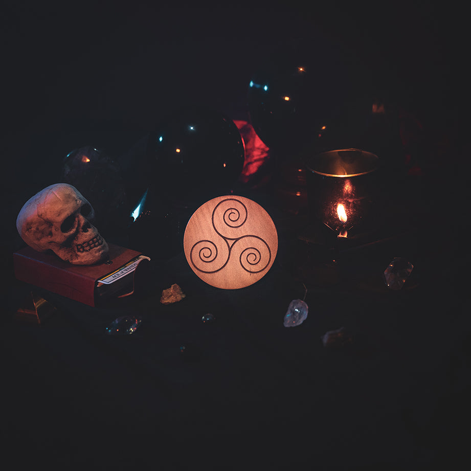 A circular wooden laser-cut crystal grid engraved with the Celtic Triskelion symbol. The grid sits amongst candles, crystal balls, a skull, tarot cards and assorted crystals.