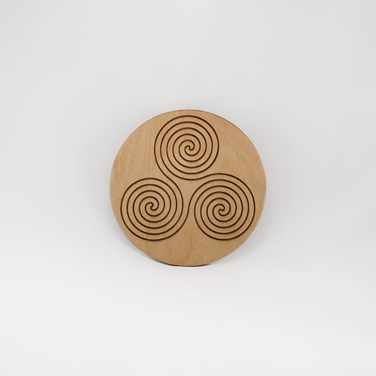 A circular wooden laser-cut crystal grid engraved with the Celtic Triskelion symbol with a plain white background.