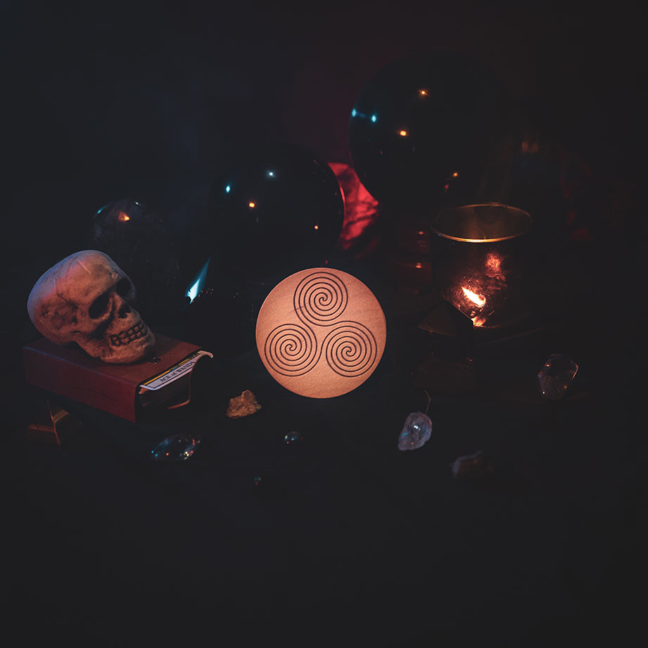 A circular wooden laser-cut crystal grid engraved with the Celtic Triskelion symbol. The grid sits amongst candles, crystal balls, a skull, tarot cards and assorted crystals.