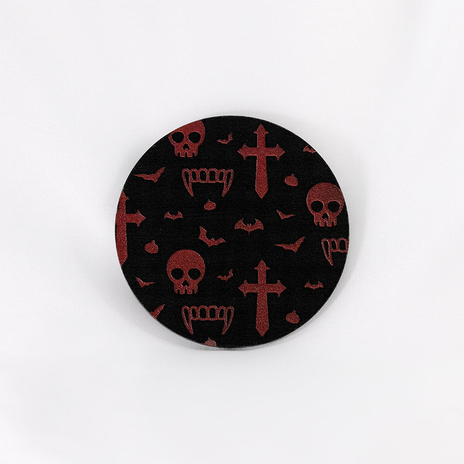A round black coaster engraved with dark red vampire symbols, including skulls, fangs, garlic, crosses and bats. The coaster is on a white background.