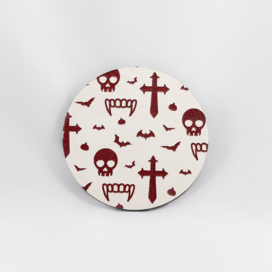 A round white coaster engraved with dark red vampire symbols, including skulls, fangs, garlic, crosses and bats. The coaster is on a white background.