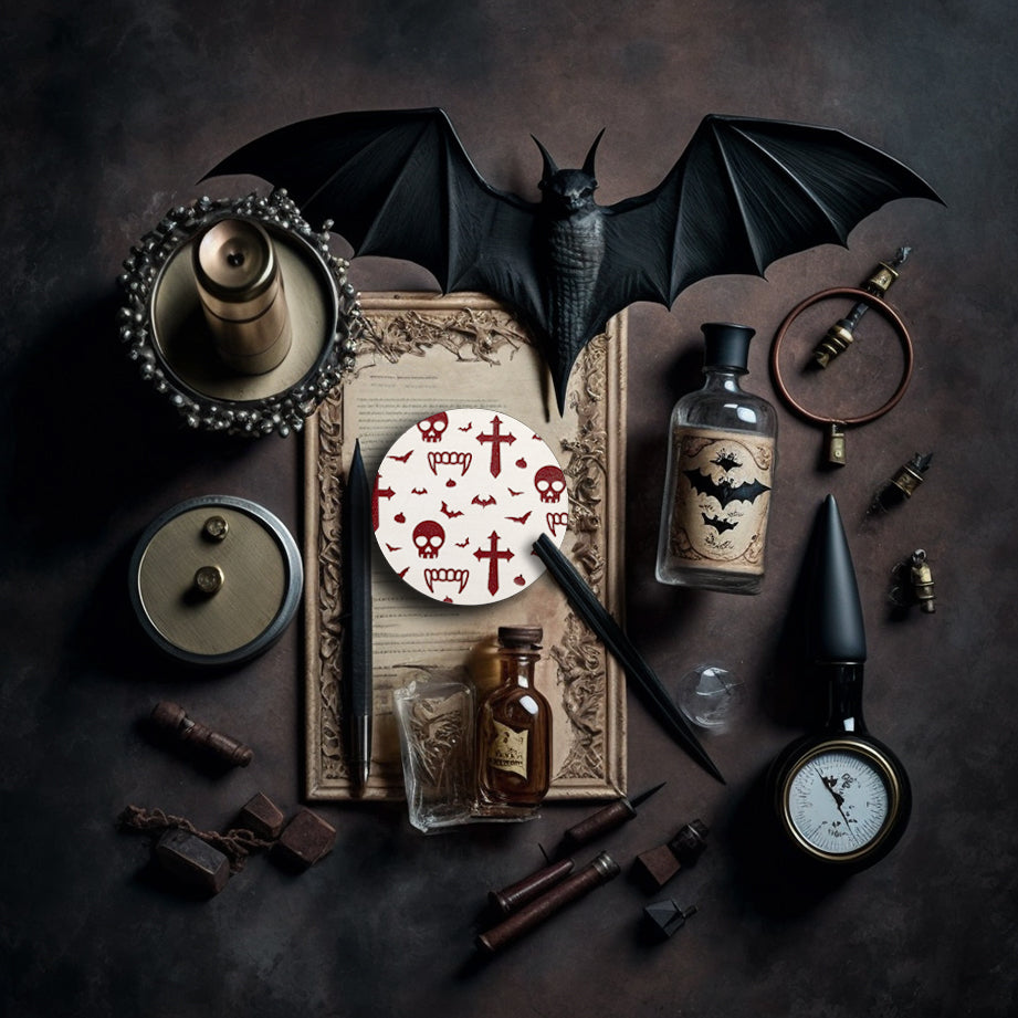 A round white coaster engraved with dark red vampire symbols, including skulls, fangs, garlic, crosses and bats. The coaster is on an old piece of paper surrounded by bats, quills, an old magnifying glass and small glass bottles.