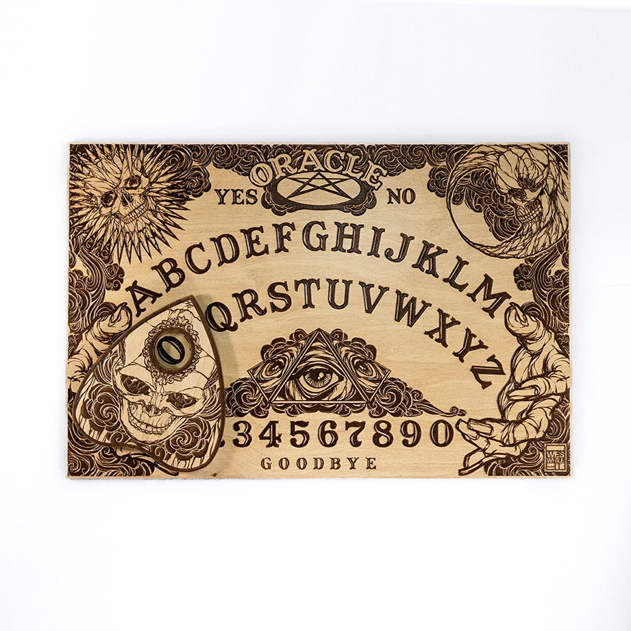 A rectangular wooden laser-cut spirit board engraved with the alphabet and numbers 0-9, yes, no, and goodbye. A sun with a skull face is engraved in the top left corner, a moon with a skull face is engraved in the top right corner, the bottom left and right corners are decorated with outstretched hands. A planchette engraved with a skull is placed on top of the board. The board is on a plain white background. 