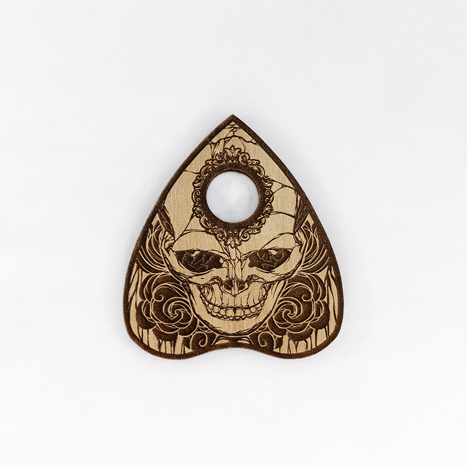 A planchette engraved with a skull on a plain white background. There is a circular hole in the forehead of the skull to see the spirit board letters through. 