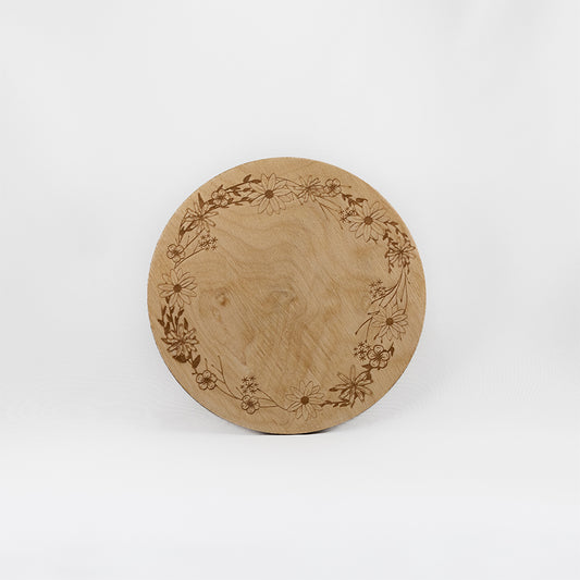 A circular wooden laser-cut crystal grid engraved with a border made up of daisies, poppies, and assorted other wildflowers and leaves with a plain white background. 