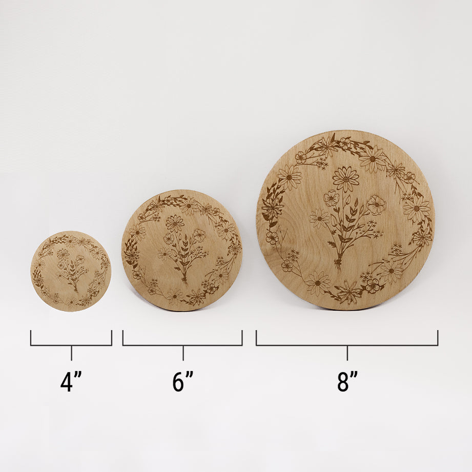 Three different sizes of the circular wooden laser-cut crystal grid engraved with a border of wildflowers and a bouquet of wildflowers in the centre against a white background with a ruler underneath all three. The smallest measuring 4", the middle 6", and the largest 8".