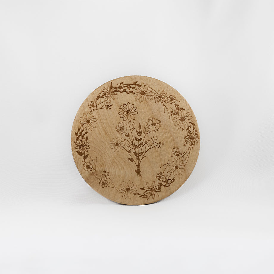 A circular wooden laser-cut crystal grid engraved with a border made up of wildflowers with a bouquet of wildflowers in the centre with a plain white background. 