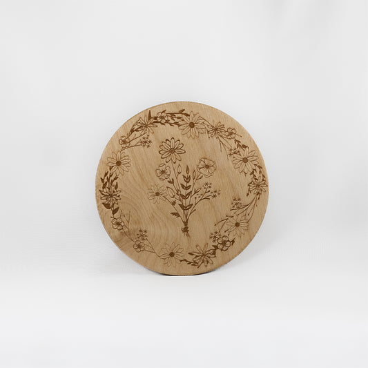 A circular wooden laser-cut crystal grid engraved with a border made up of wildflowers with a bouquet of wildflowers in the centre with a plain white background. 