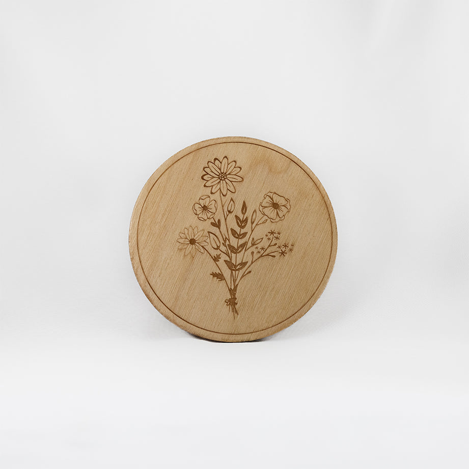 A circular wooden laser-cut crystal grid engraved in the centre with a bouquet of wildflowers with a plain white background. 